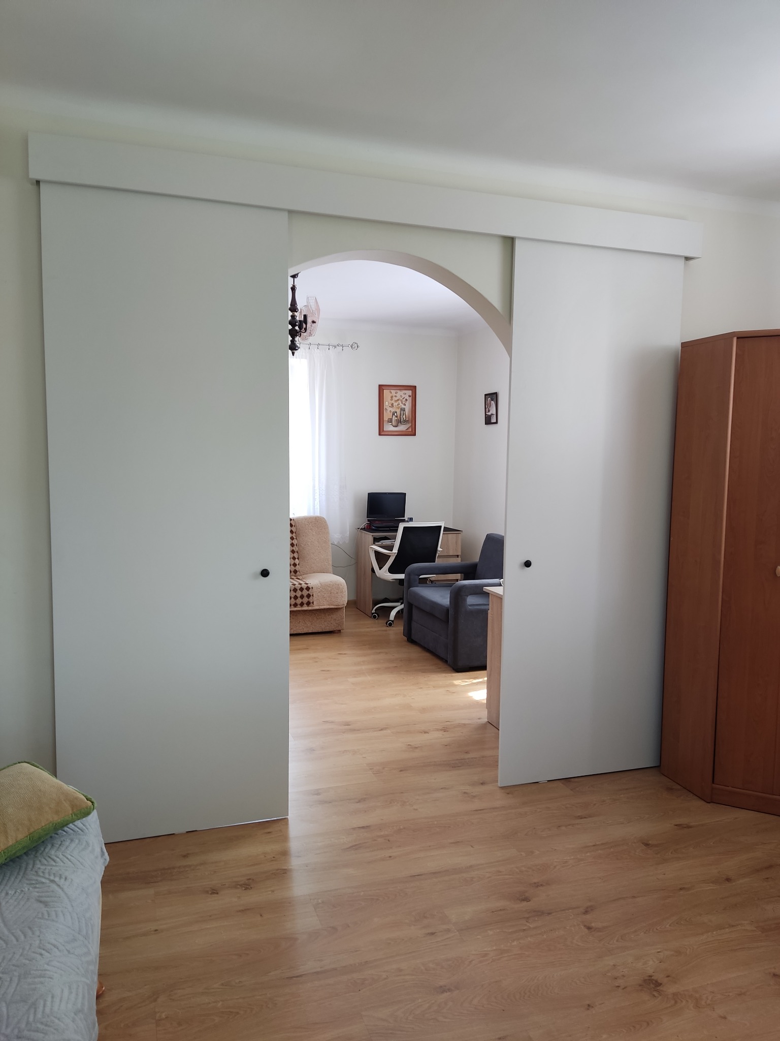 Białe, przesuwne drzwi wnękowe z łukowym przejściem do pokoju w Rębowie. Widoczne drewniane panele podłogowe i meble w tle. Minimalistyczny design.
