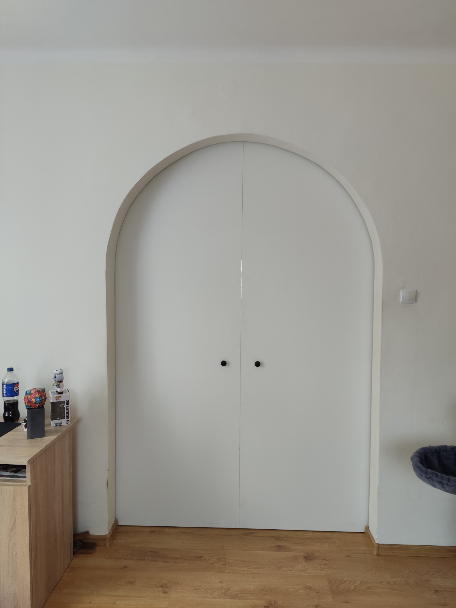 Białe, dwuskrzydłowe drzwi wnękowe z zaokrąglonym u góry otworem, z czarnymi, okrągłymi uchwytami, osadzone w białej ścianie. Widoczny fragment podłogi i wyposażenia wnętrza.