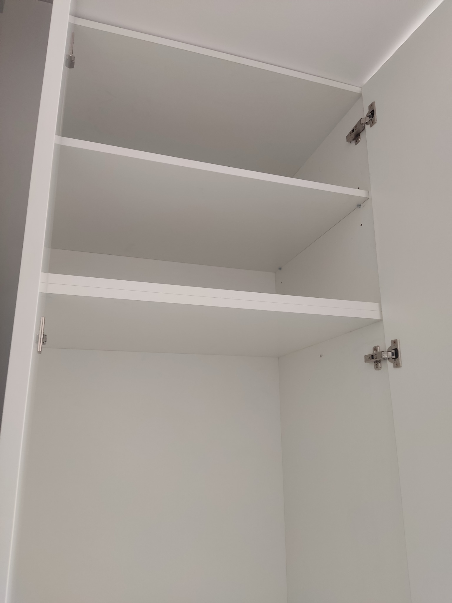 Wnętrze białej szafy wnękowej z trzema półkami, widoczne zawiasy i mocowania. Ujęcie od dołu, podkreślające przestrzeń i symetrię półek.