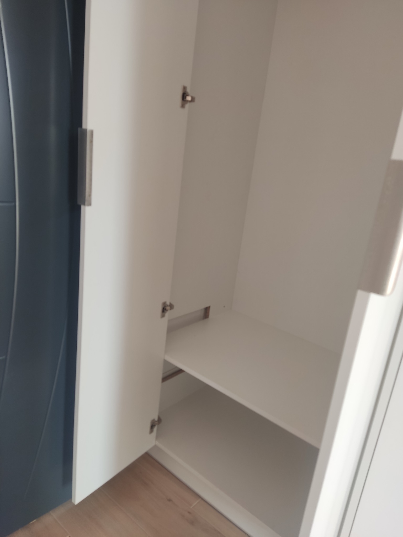 Wnętrze białej szafy z półkami, widoczne zawiasy i metalowy uchwyt na drzwiach. Drzwi szafy częściowo otwarte, ukazujące strukturę półek wewnątrz.
