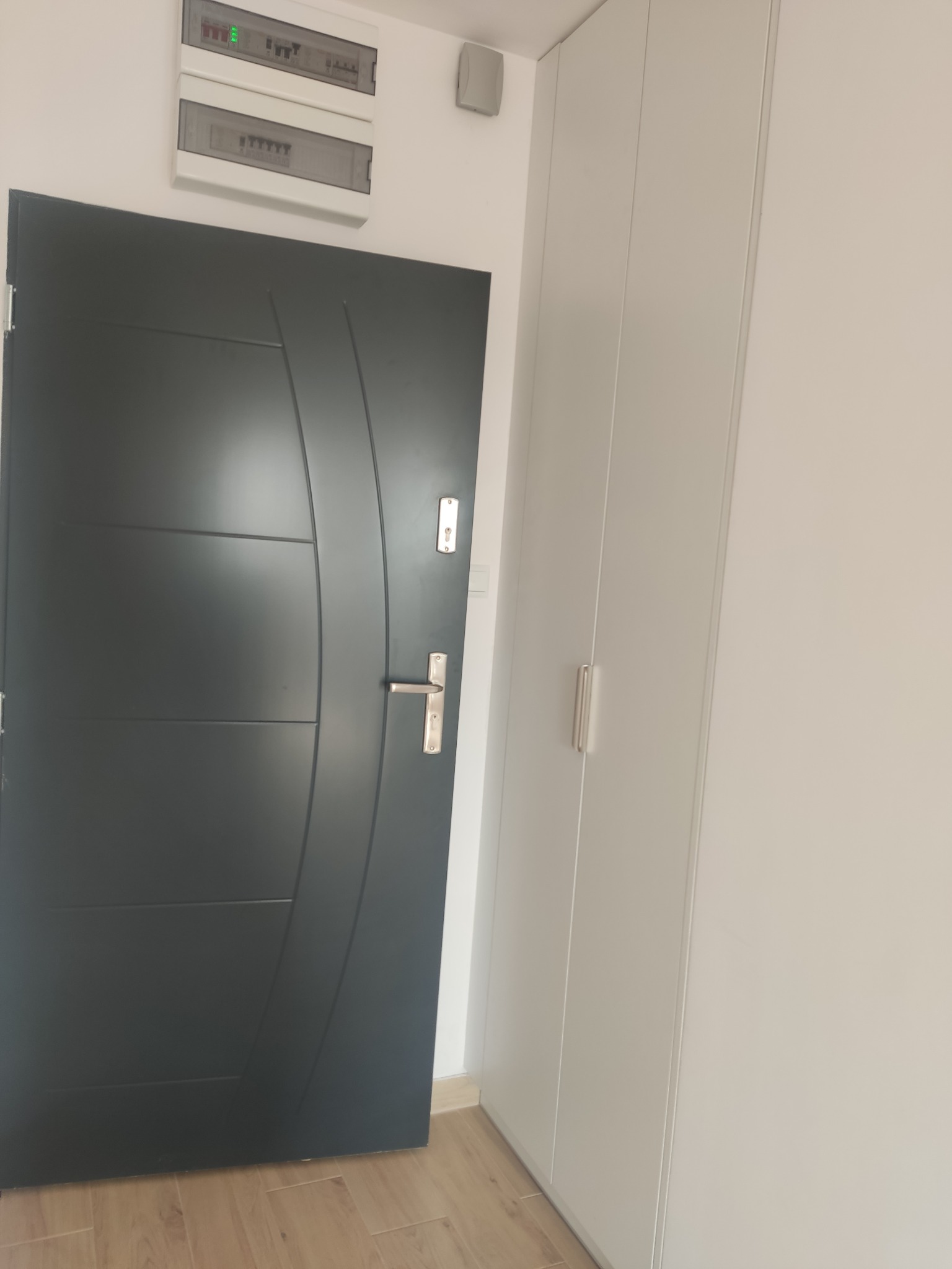 Szara, nowoczesna szafa wnękowa z drzwiami przesuwnymi, zamontowana obok wejściowych drzwi. Widoczna skrzynka z bezpiecznikami.
