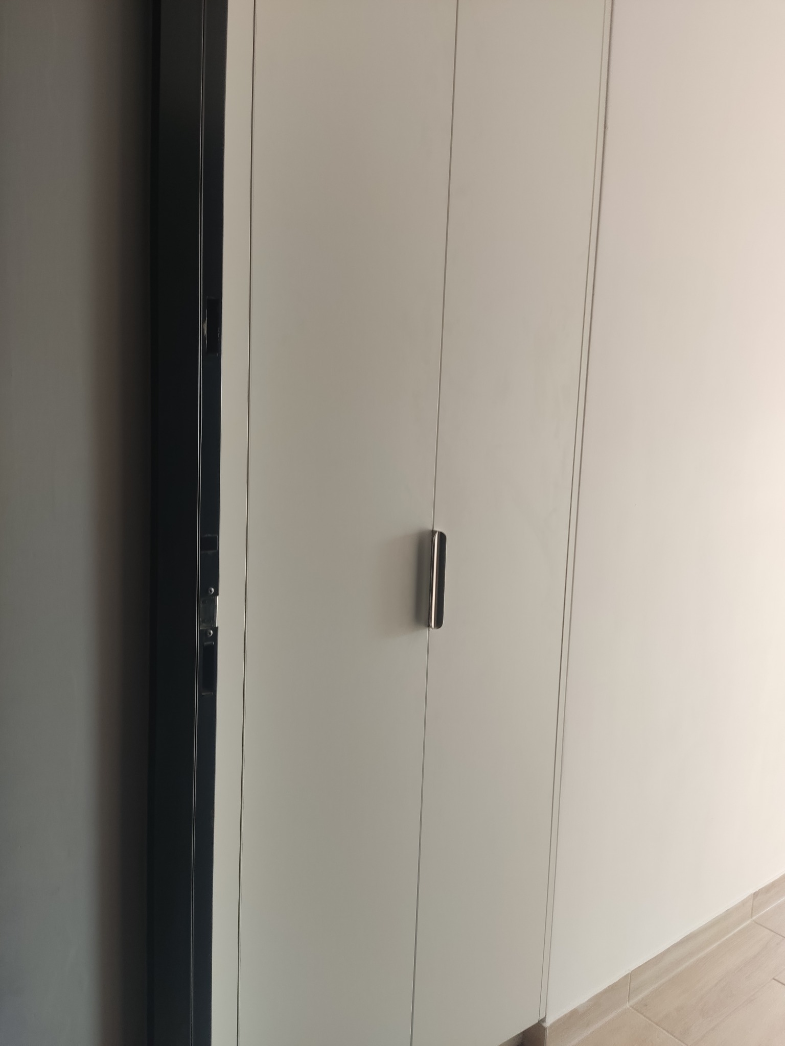Zamknięta, biała szafa wnękowa z minimalistycznym, srebrnym uchwytem na tle jasnej ściany i fragmentu podłogi z płytek. Widoczny fragment ramy drzwi.