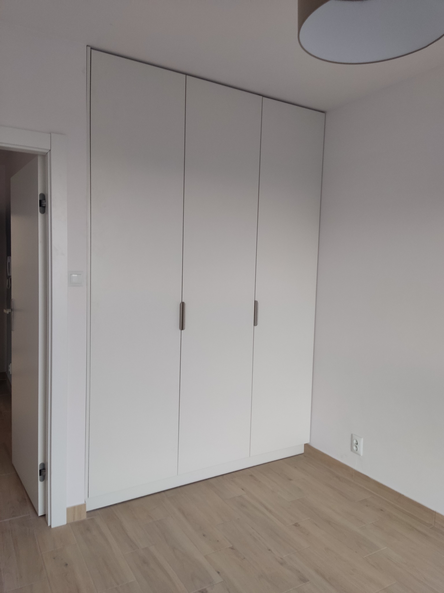 Minimalistyczna, biała szafa wnękowa z trzema drzwiami i subtelnymi uchwytami, w jasnym wnętrzu z drewnopodobną podłogą. Widoczny fragment lampy sufitowej.
