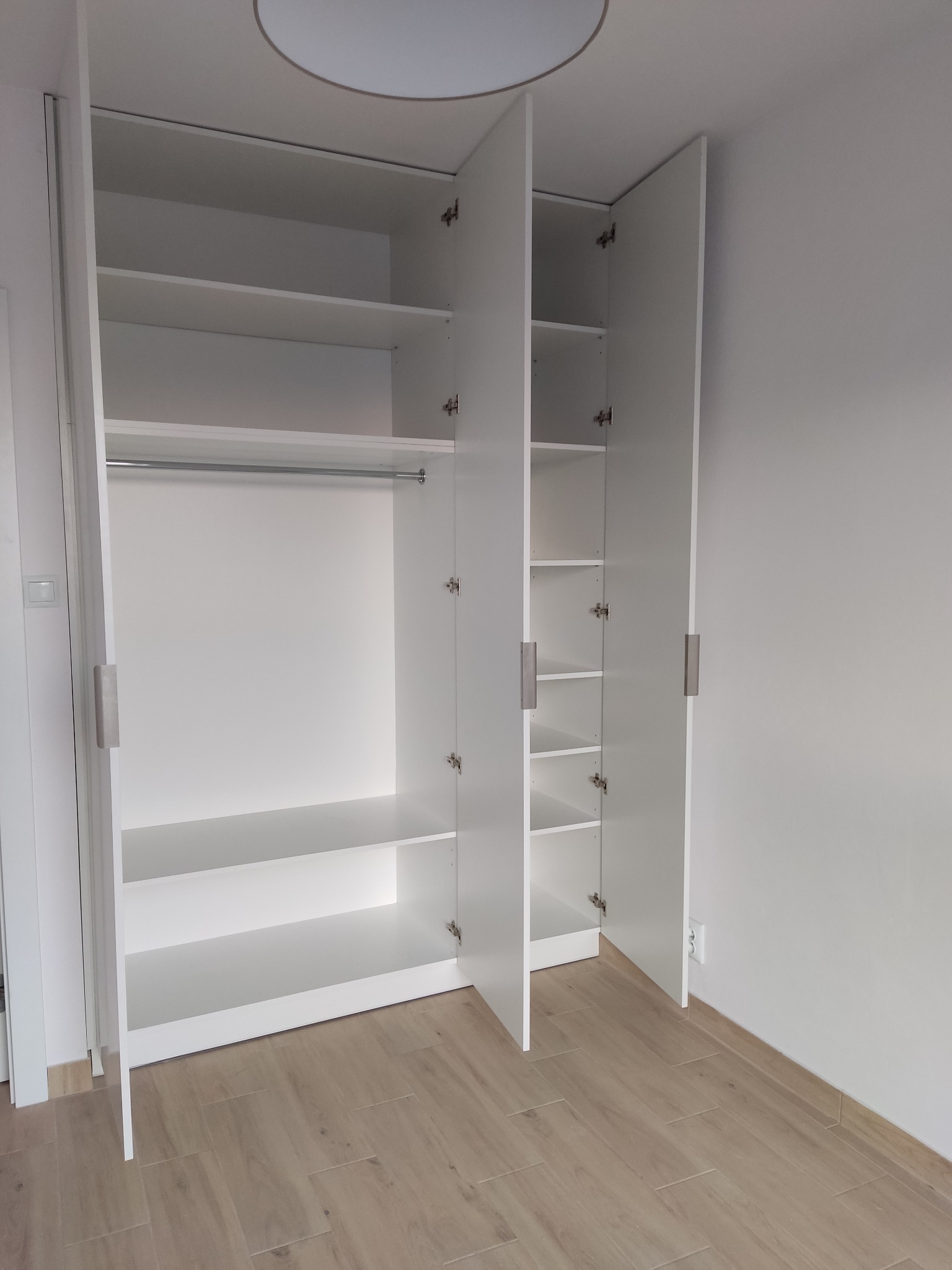 Biała szafa wnękowa z otwartymi drzwiami, ukazująca półki i drążek na ubrania. Widoczna drewnopodobna podłoga i minimalistyczne oświetlenie sufitowe.