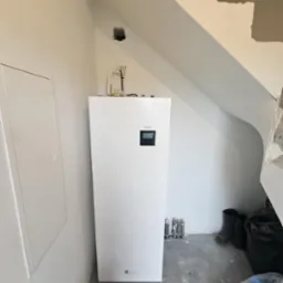 Biała pompa ciepła Midea zamontowana w rogu pomieszczenia ze skośnym sufitem, widoczne rury i otwór w ścianie, podłoga w trakcie prac wykończeniowych.