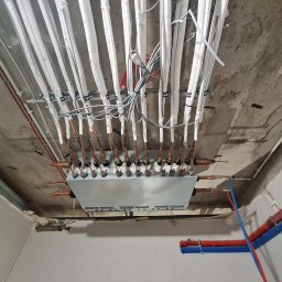KLASS-INSTAL Szymon Klass - Rozdzielacz instalacji chłodniczej z miedzianymi rurkami i izolowanymi przewodami przymocowany do surowego betonowego sufitu, widoczne również rury instalacyjne w kolorze czerwonym i niebieskim.