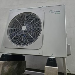 KLASS-INSTAL Szymon Klass - Biała jednostka zewnętrzna klimatyzacji Midea R32 zamontowana na betonowych bloczkach z czarnymi gumowymi podkładkami antywibracyjnymi, widoczna na tle szarej ściany budynku.