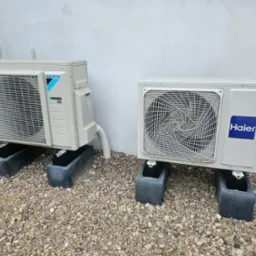 Dwa klimatyzatory zewnętrzne, marki Daikin i Haier, ustawione na betonowych podstawach na żwirowym podłożu, przy jasnej ścianie budynku i ogrodzeniu z siatki.