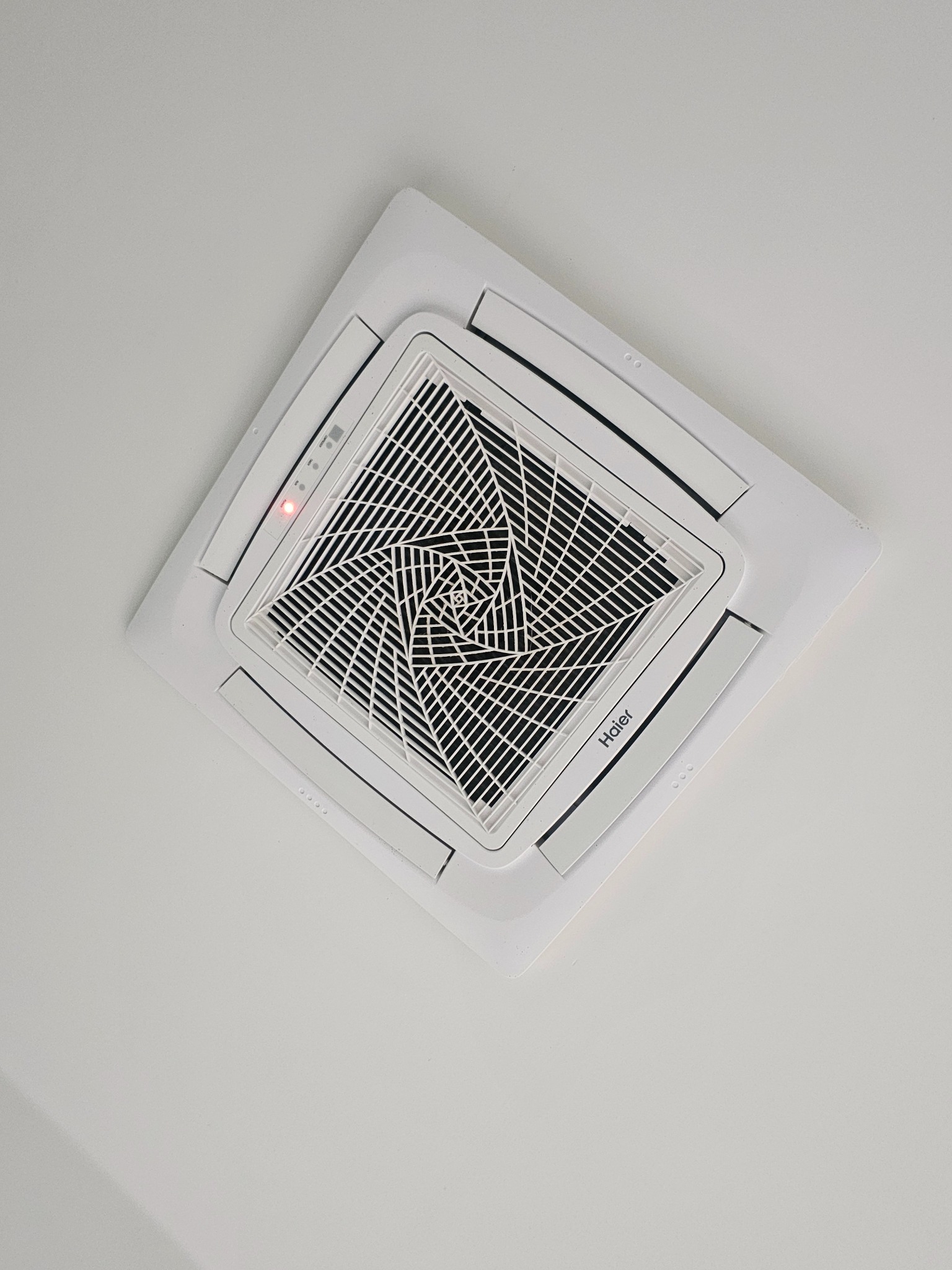 Biała, sufitowa kaseta klimatyzacyjna marki Haier, z widoczną kratką wentylacyjną i czerwoną diodą kontrolną, zamontowana na białym suficie, ujęcie pod kątem.