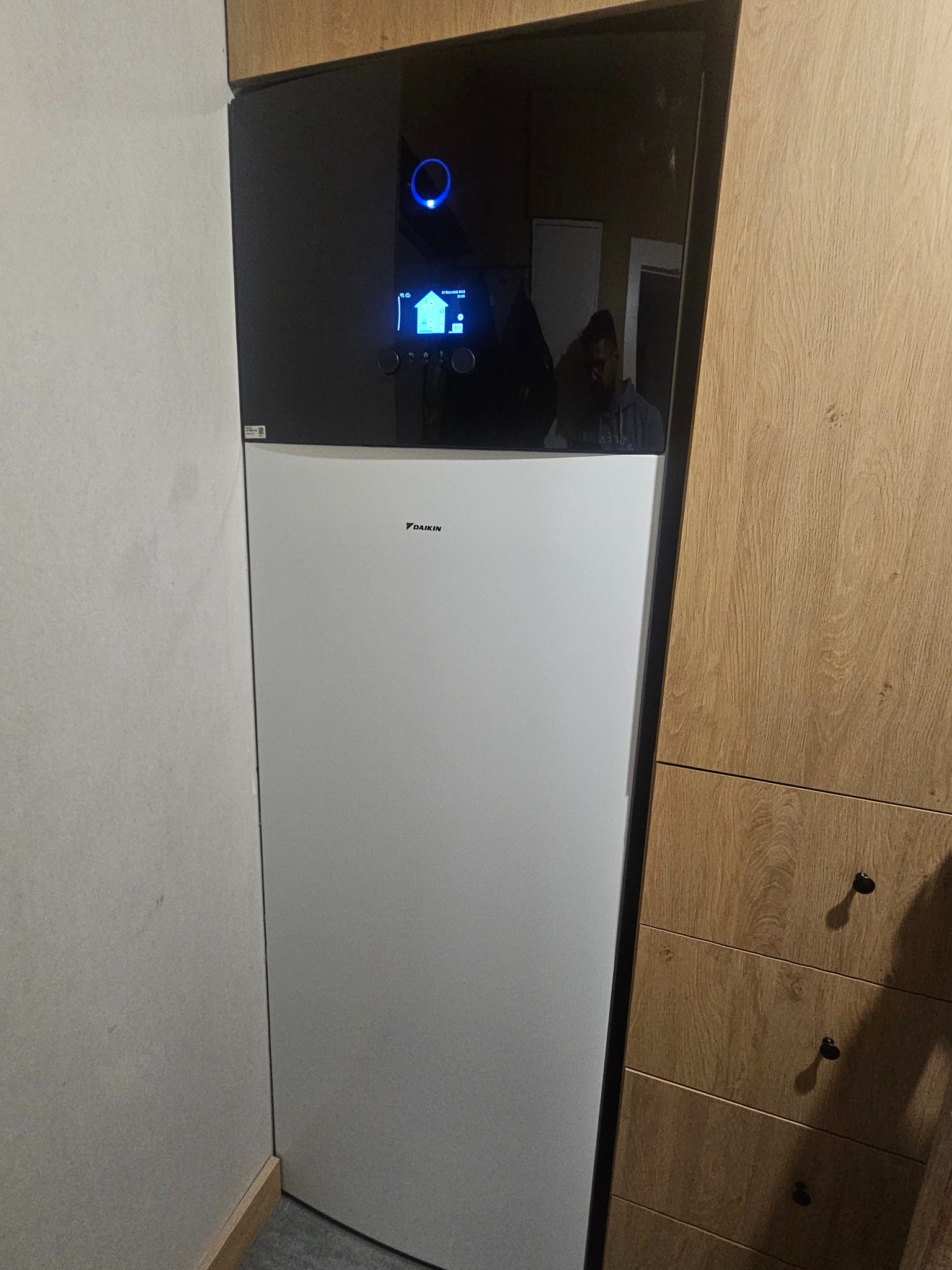 Biała pompa ciepła Daikin zamontowana w rogu pomieszczenia, widoczny wyświetlacz z niebieskim podświetleniem oraz fragment drewnianej komody. W odbiciu na górnej części obudowy widać sylwetkę osoby.