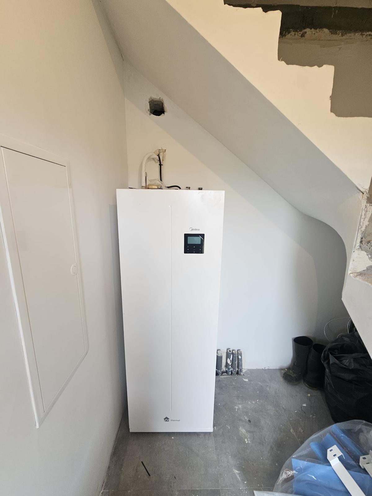 Biała pompa ciepła Midea zamontowana w rogu pomieszczenia ze skośnym sufitem, widoczne rury i otwór w ścianie, podłoga w trakcie prac wykończeniowych.