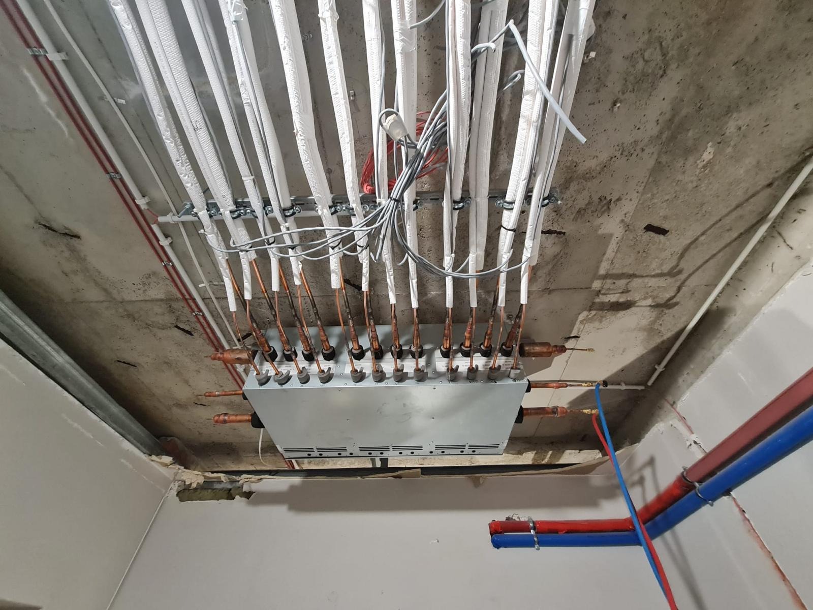 Rozdzielacz instalacji chłodniczej z miedzianymi rurkami i izolowanymi przewodami przymocowany do surowego betonowego sufitu, widoczne również rury instalacyjne w kolorze czerwonym i niebieskim.