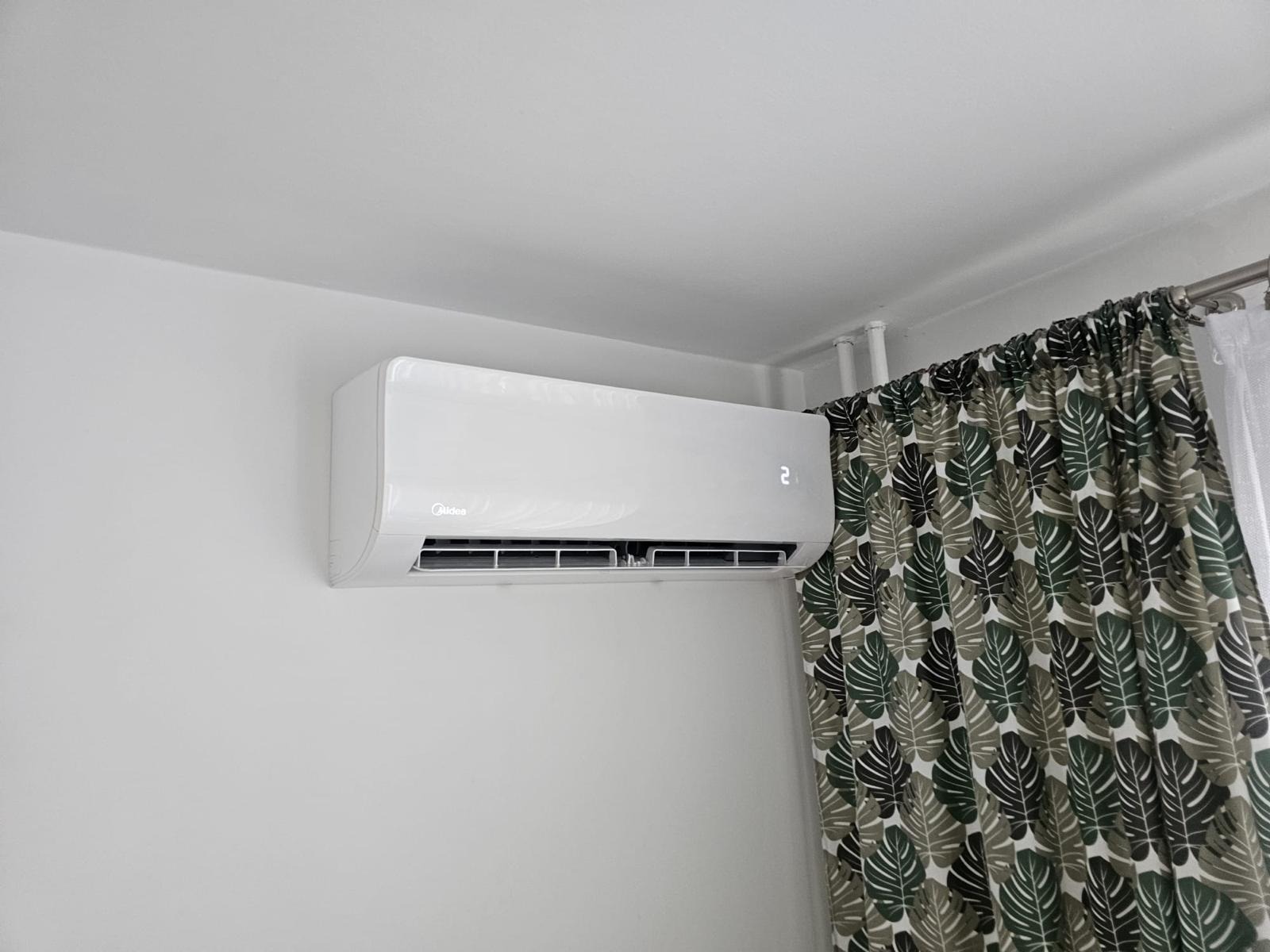 Biały klimatyzator Midea zamontowany na ścianie w pomieszczeniu z zasłoną w liście.