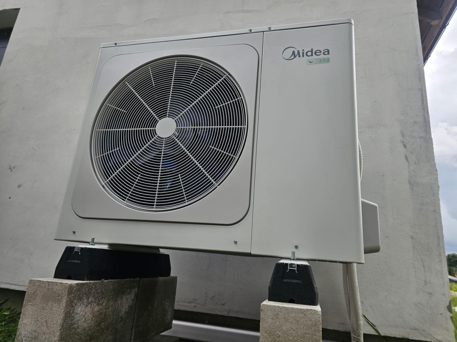 Biała jednostka zewnętrzna klimatyzacji Midea R32 zamontowana na betonowych bloczkach z czarnymi gumowymi podkładkami antywibracyjnymi, widoczna na tle szarej ściany budynku.