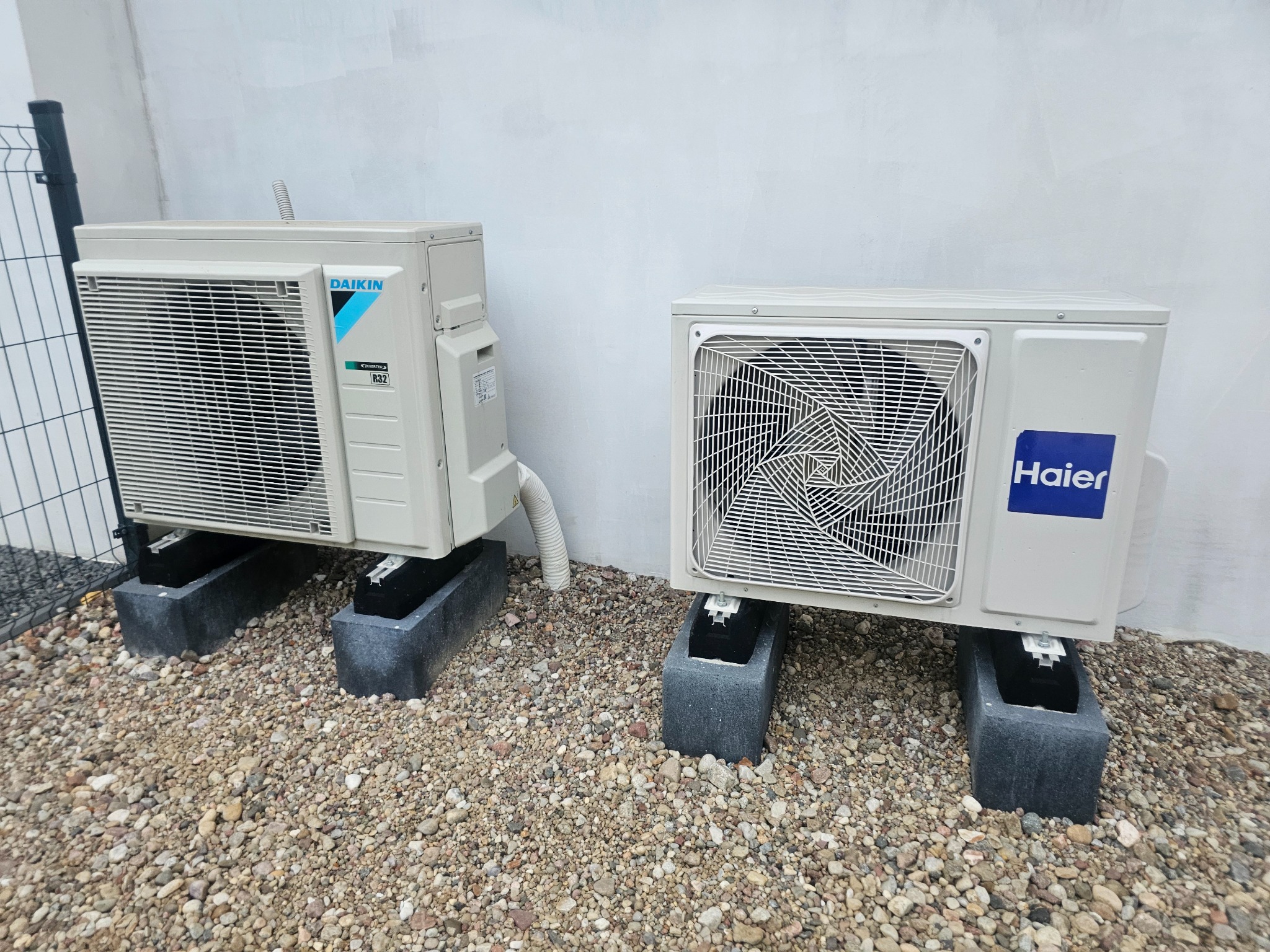 Dwa klimatyzatory zewnętrzne, marki Daikin i Haier, ustawione na betonowych podstawach na żwirowym podłożu, przy jasnej ścianie budynku i ogrodzeniu z siatki.
