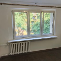 ArtBud - Białe, trzyskrzydłowe okno PCV z widokiem na zieleń drzew i blok z balkonami, zamontowane nad białym, żeliwnym grzejnikiem w pokoju z parkietem ułożonym w jodełkę.