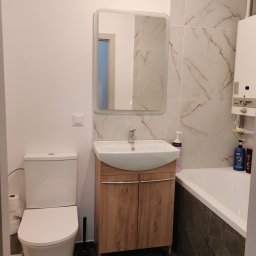 ArtBud - Nowoczesna łazienka z umywalką w szafce z drewnopodobnym frontem, lustrem z podświetleniem LED, toaletą, wanną obudowaną ciemnymi płytkami oraz grzejnikiem gazowym na ścianie z jasnymi płytkami...