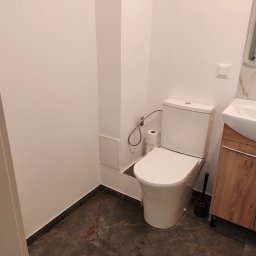 ArtBud - Nowoczesna toaleta z białą ceramiką, marmurową podłogą i drewnianą szafką pod umywalką, widoczna rolka papieru toaletowego i szczotka do WC.