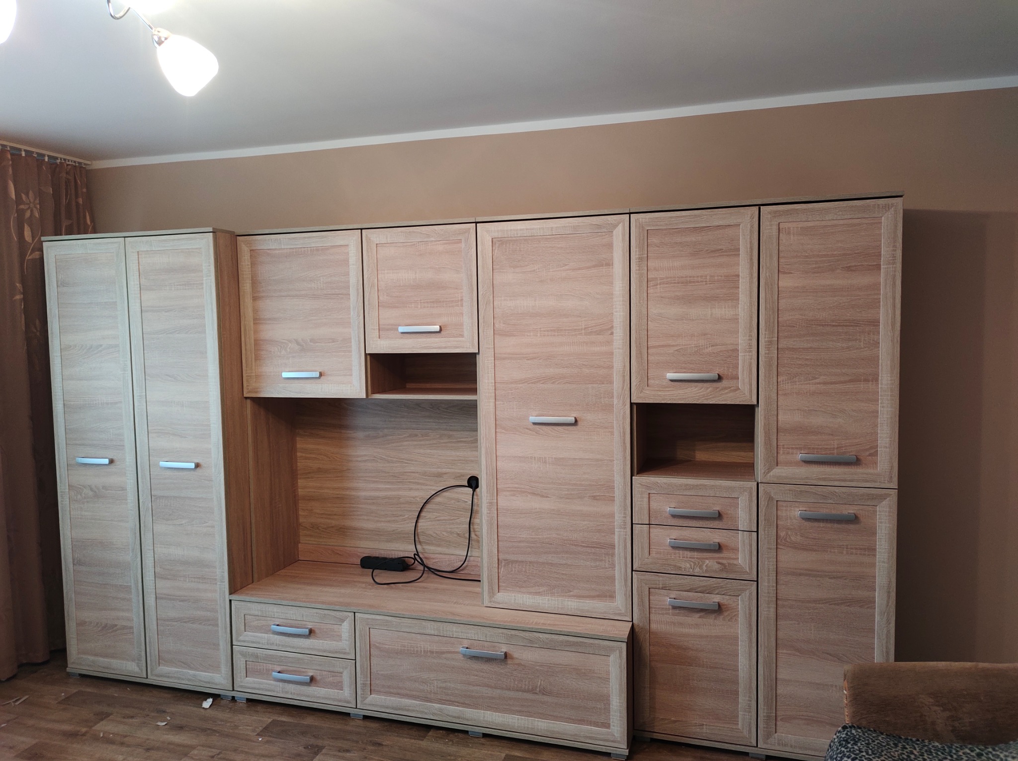 Kompletna, jasna meblościanka z drewnopodobnym wykończeniem, z szafkami, szufladami i otwartą przestrzenią, na tle beżowej ściany i fragmentu zasłony.