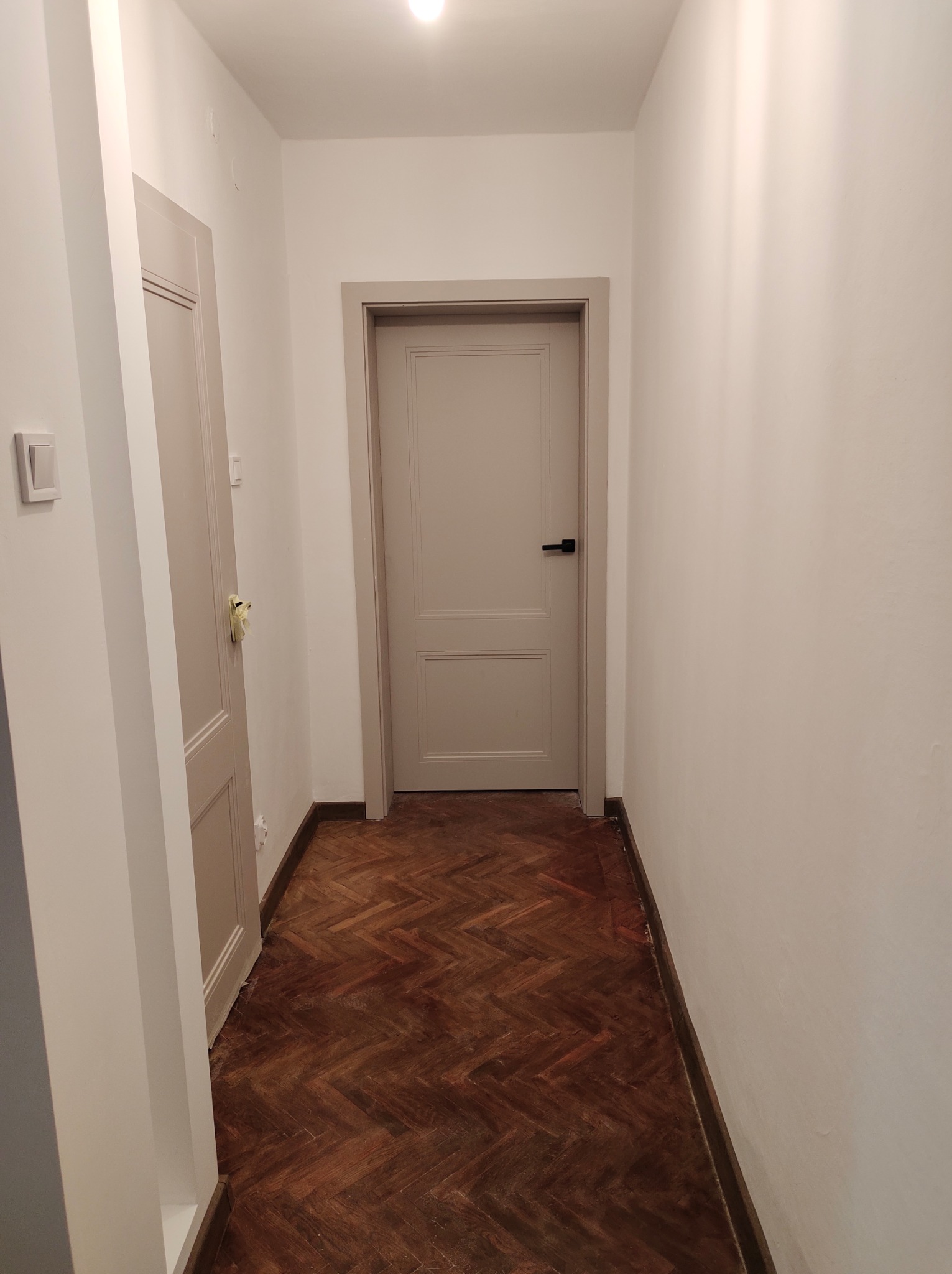 Korytarz po odświeżeniu: białe ściany, odnowiona drewniana podłoga w jodełkę, dwoje drzwi w odcieniach szarości z czarnymi klamkami, widoczny włącznik światła.