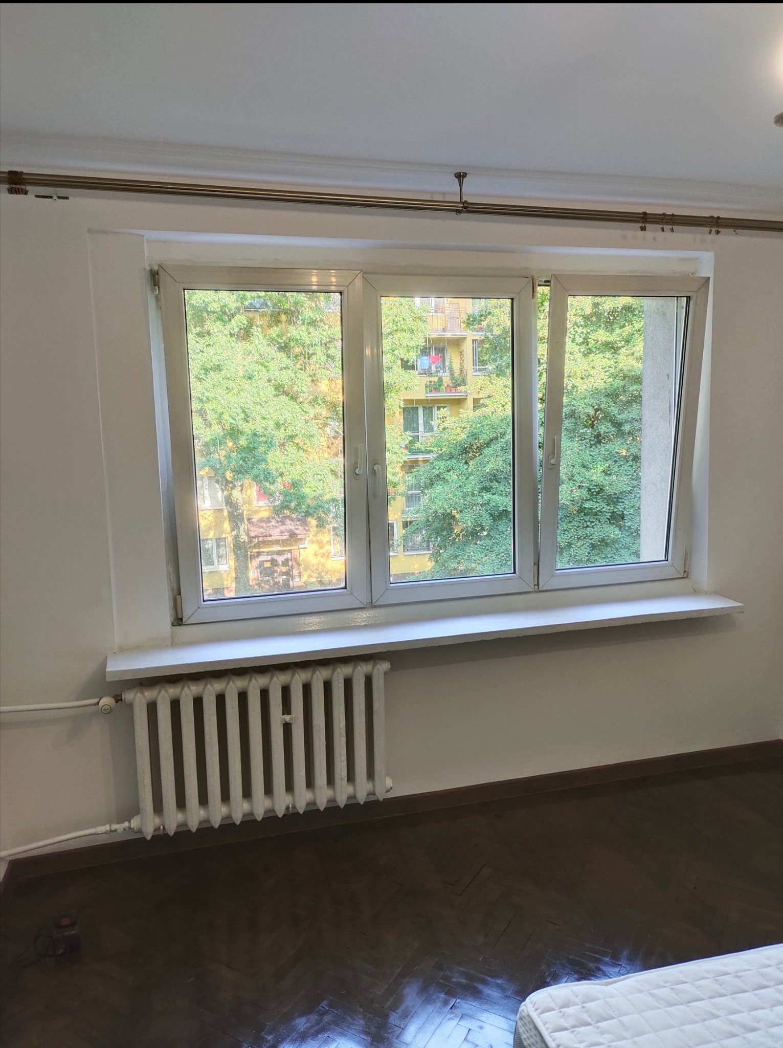 Białe, trzyskrzydłowe okno PCV z widokiem na zieleń drzew i blok z balkonami, zamontowane nad białym, żeliwnym grzejnikiem w pokoju z parkietem ułożonym w jodełkę.