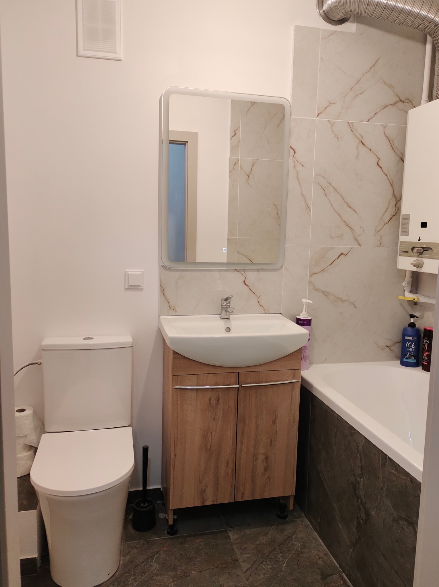 Nowoczesna łazienka z umywalką w szafce z drewnopodobnym frontem, lustrem z podświetleniem LED, toaletą, wanną obudowaną ciemnymi płytkami oraz grzejnikiem gazowym na ścianie z jasnymi płytkami...