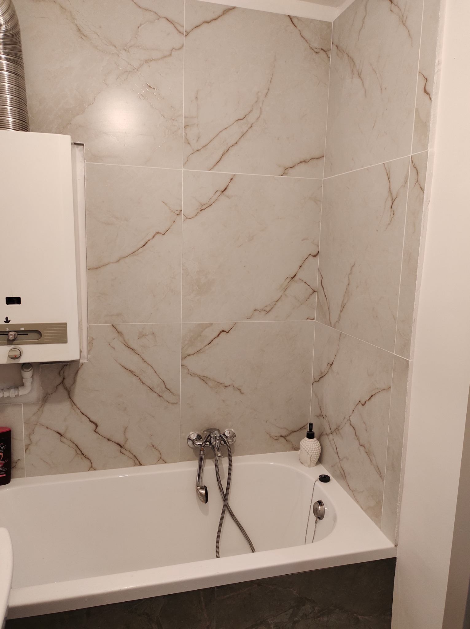 Wykończona łazienka z wanną, obudowana płytkami imitującymi marmur, widoczny bojler gazowy i armatura.