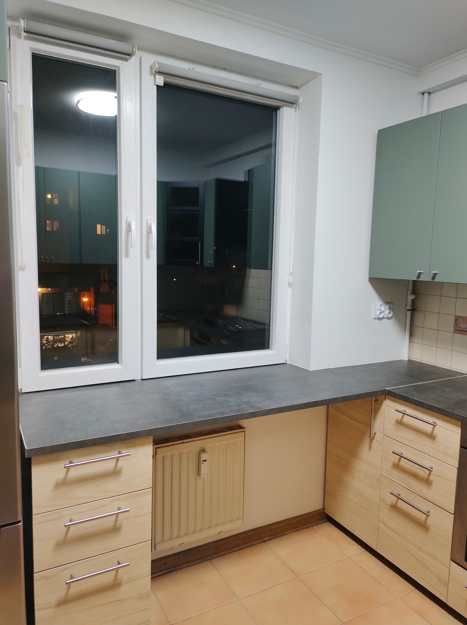 Fragment kuchni z oknem: szare blaty, jasne drewniane szafki z metalowymi uchwytami, zielone szafki wiszące, terakotowa podłoga. Widok na nocne miasto za oknem.