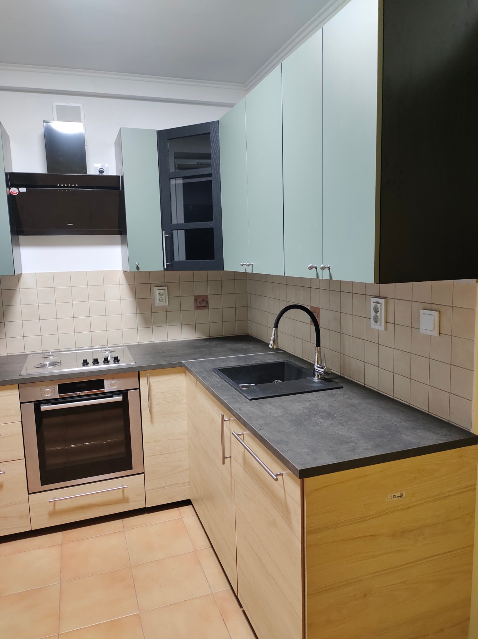 Nowoczesna kuchnia z jasnymi drewnianymi szafkami dolnymi, pastelowymi górnymi i czarnymi akcentami, w tym zlew i okap; beżowe płytki na ścianie i brązowe na podłodze.