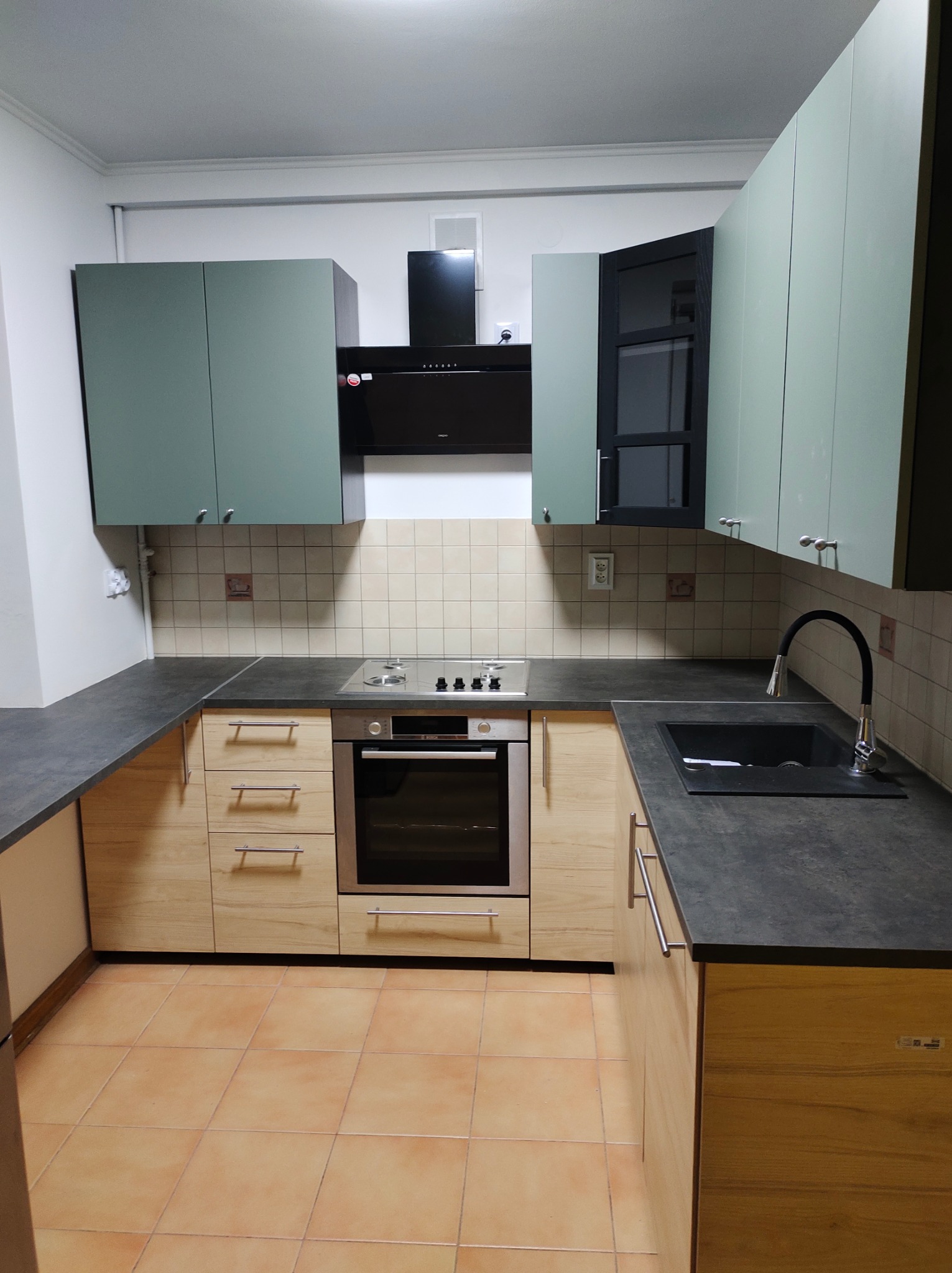 Wykończona kuchnia z drewnianymi szafkami dolnymi i zielonymi górnymi, czarnym blatem, płytą grzewczą i piekarnikiem, oraz czarnym okapem i zlewem, płytki na podłodze w kolorze terakoty.