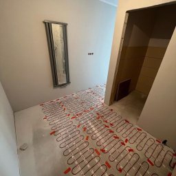 Sowrem - Wykończenia wnętrz Rybnik - Rozłożona mata grzewcza na wylewce w korytarzu, przygotowana do dalszych prac wykończeniowych. Na ścianie wisi wąskie lustro.