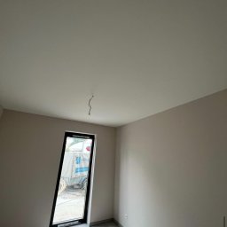 Sowrem - Wykończenia wnętrz Rybnik - Wnętrze pokoju po szpachlowaniu ścian na gładko, widoczne okno z czarną ramą i wystający kabel ze ściany. Na zewnątrz przyczepa z logo firmy.
