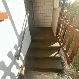Sowrem - Wykończenia wnętrz Rybnik - Świeżo wylane betonowe schody wejściowe z metalową poręczą przy białej ścianie domu, widoczne płytki na ścianie werandy i fragment drewnianego płotku.