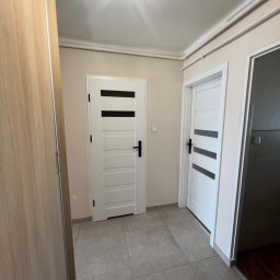 Sowrem - Wykończenia wnętrz Rybnik - Korytarz z dwiema białymi, nowoczesnymi drzwiami z czarnymi poziomymi paskami, szara podłoga z płytek, jasne ściany i drewniana szafa w tle.