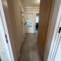 Sowrem - Wykończenia wnętrz Rybnik - Wąski korytarz z trzema białymi drzwiami o różnych wzorach klamek i przeszkleń, wyłożony szarą, kwadratową terakotą, z widoczną tapetą w kwiatowy wzór po lewej stronie.