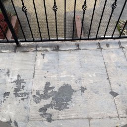 Sowrem - Wykończenia wnętrz Rybnik - Przygotowanie podłoża na balkonie pod hydroizolację, widoczne ślady po szpachlowaniu i usuniętych starych płytkach, balustrada metalowa z ozdobnymi elementami w tle bruk na ulicy.