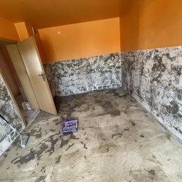 Sowrem - Wykończenia wnętrz Rybnik - Pomarańczowe ściany w pokoju w trakcie przygotowywania podłoża, ściany częściowo pokryte warstwą wyrównującą, na podłodze ślady zaprawy, widoczny wałek i kuweta malarska.