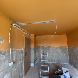 Sowrem - Wykończenia wnętrz Rybnik - Pokój w trakcie prac przygotowawczych: pomarańczowa góra ścian i sufit, dolna część ścian w szarym tynku, widoczne przewody elektryczne, drabina aluminiowa i worki z materiałami budowlanymi...