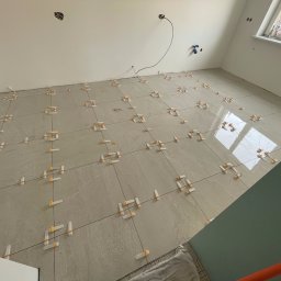 Sowrem - Wykończenia wnętrz Rybnik - Jasnoszare płytki podłogowe w trakcie układania, z widocznymi krzyżykami dystansowymi, kabel wystający ze ściany, fragment okna z widokiem na zewnątrz.