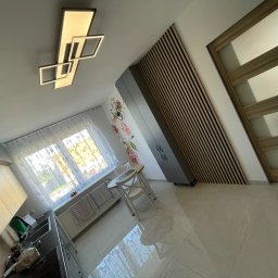 Sowrem - Wykończenia wnętrz Rybnik - Nowoczesna kuchnia z drewnianą ścianą, wiszącą lampą w geometrycznym stylu i białymi płytkami podłogowymi. Widoczny fragment blatu kuchennego, okno z firanką oraz stół z krzesłami.