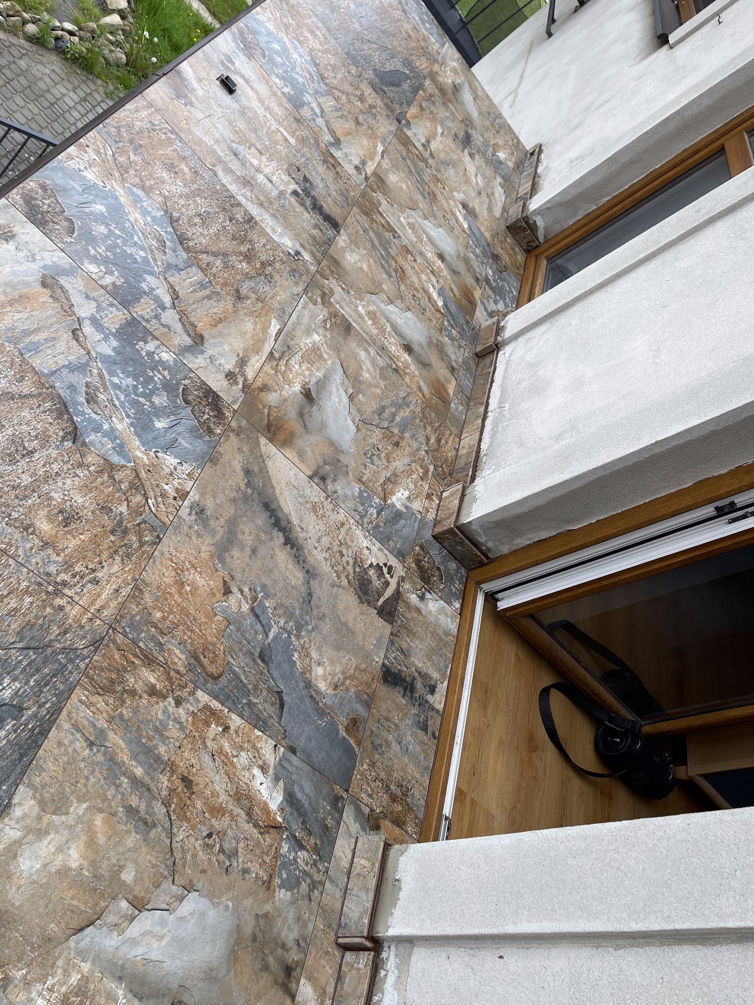 Elewacja budynku wykończona płytkami imitującymi kamień naturalny w odcieniach szarości, brązu i beżu, widoczne okno z drewnianą ramą i fragment wnętrza z głośnikiem przenośnym.