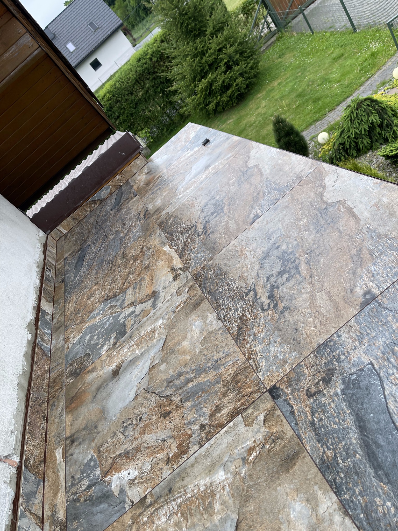 Ukończona ściana zewnętrzna wyłożona płytkami imitującymi kamień w odcieniach szarości i brązu, widok z góry na tle zieleni ogrodu i fragmentu dachu.