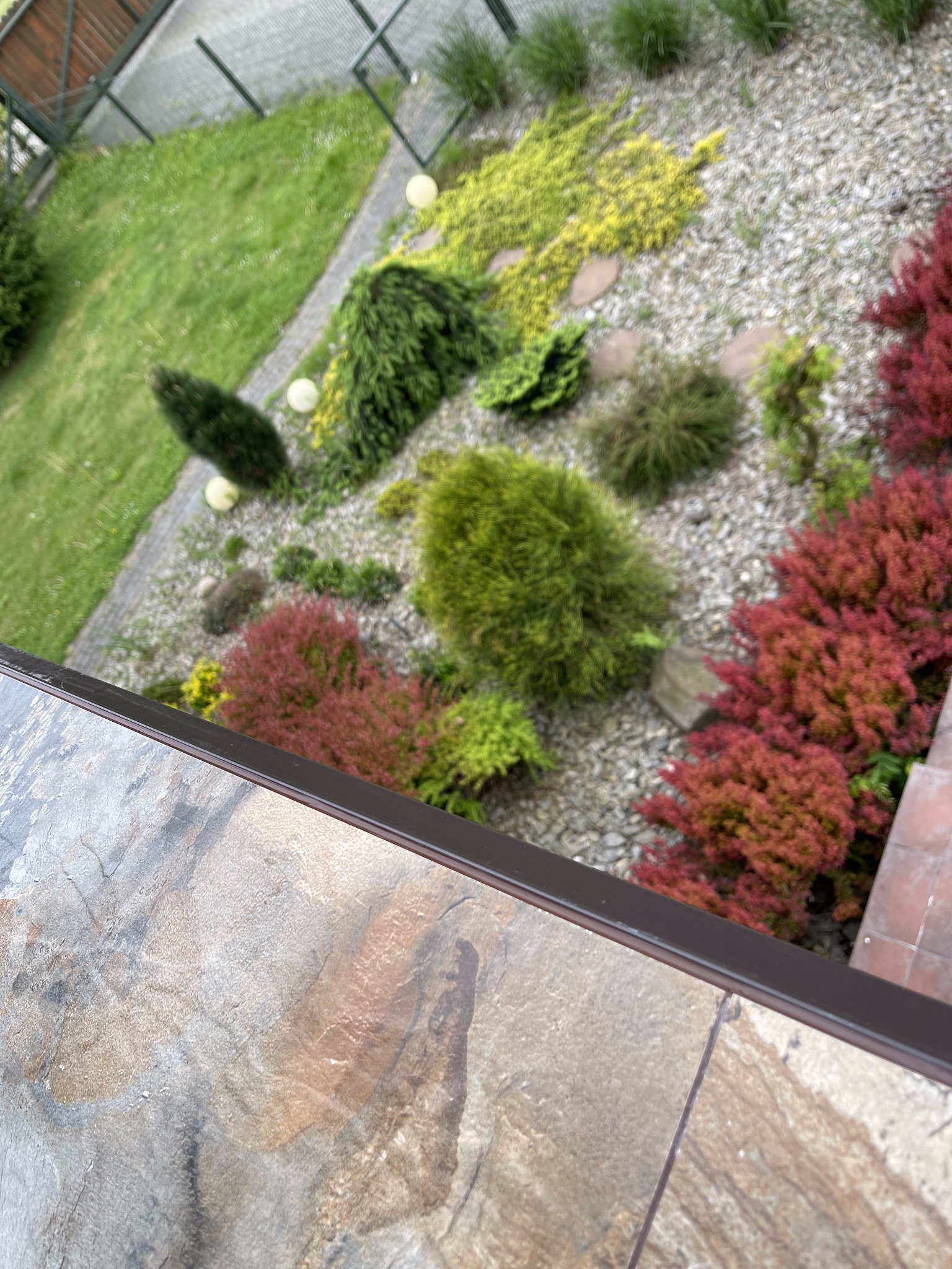 Widok z góry na fragment ogrodu z różnorodną roślinnością ozdobną, kamieniami i oświetleniem, widziany z balkonu wykończonego płytkami imitującymi kamień.