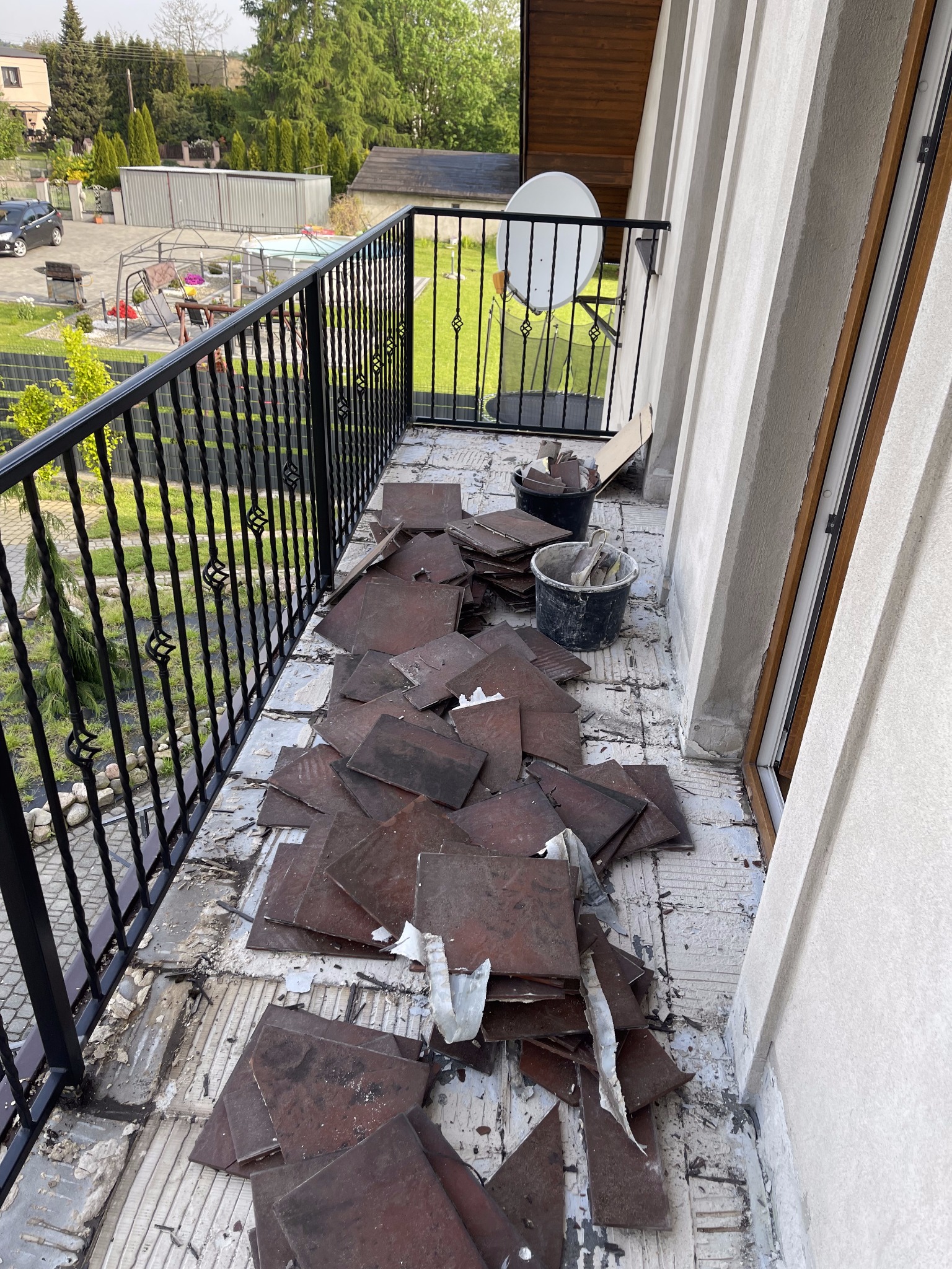 Demontaż starej terakoty na balkonie z widocznymi pozostałościami kleju i fragmentami płytek, dwa wiadra z narzędziami i resztkami płytek w tle, czarna metalowa balustrada z ozdobnymi elementami...