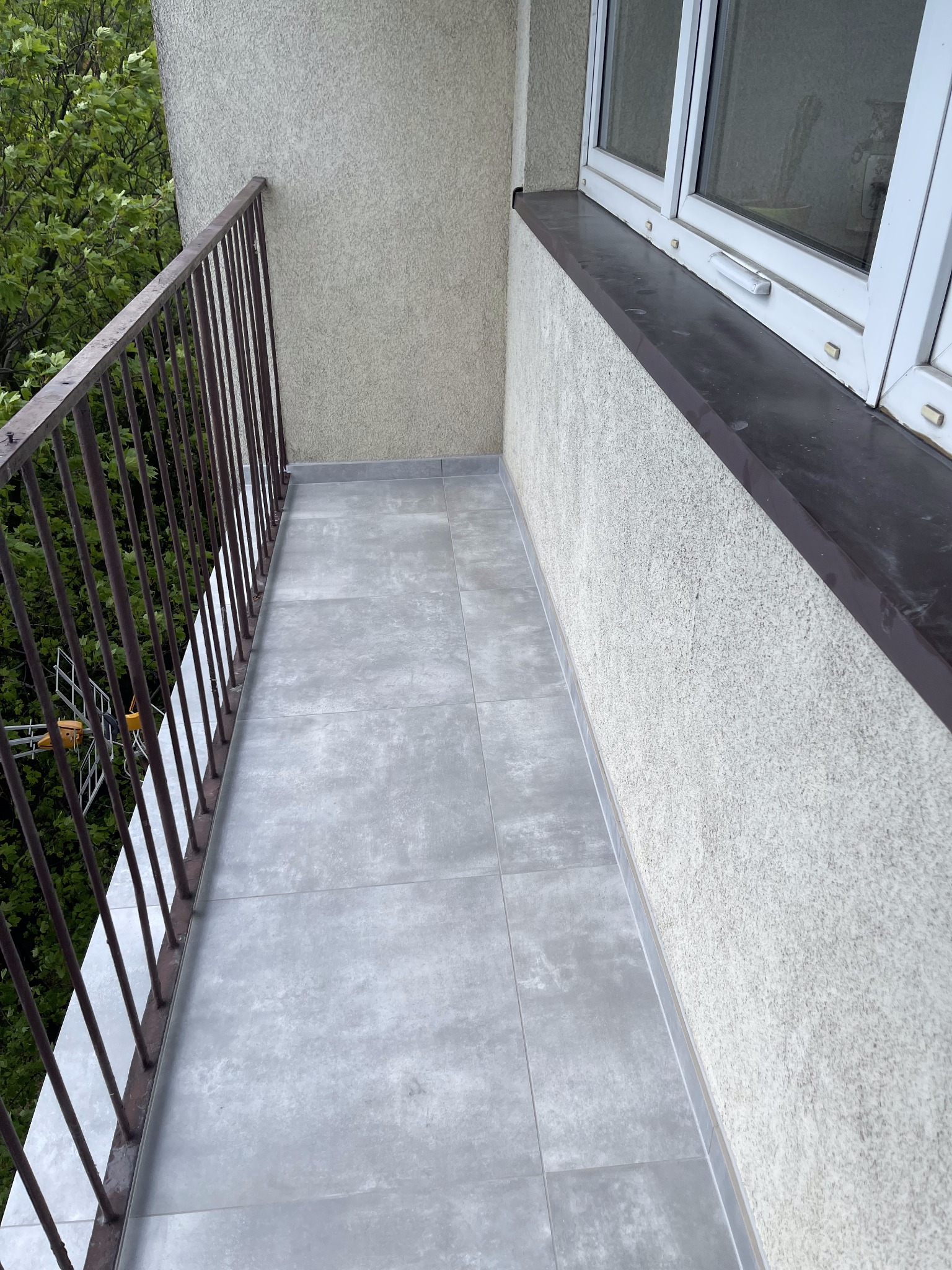 Wykończony balkon z szarymi płytkami podłogowymi, brązową balustradą i widokiem na zielone drzewa.