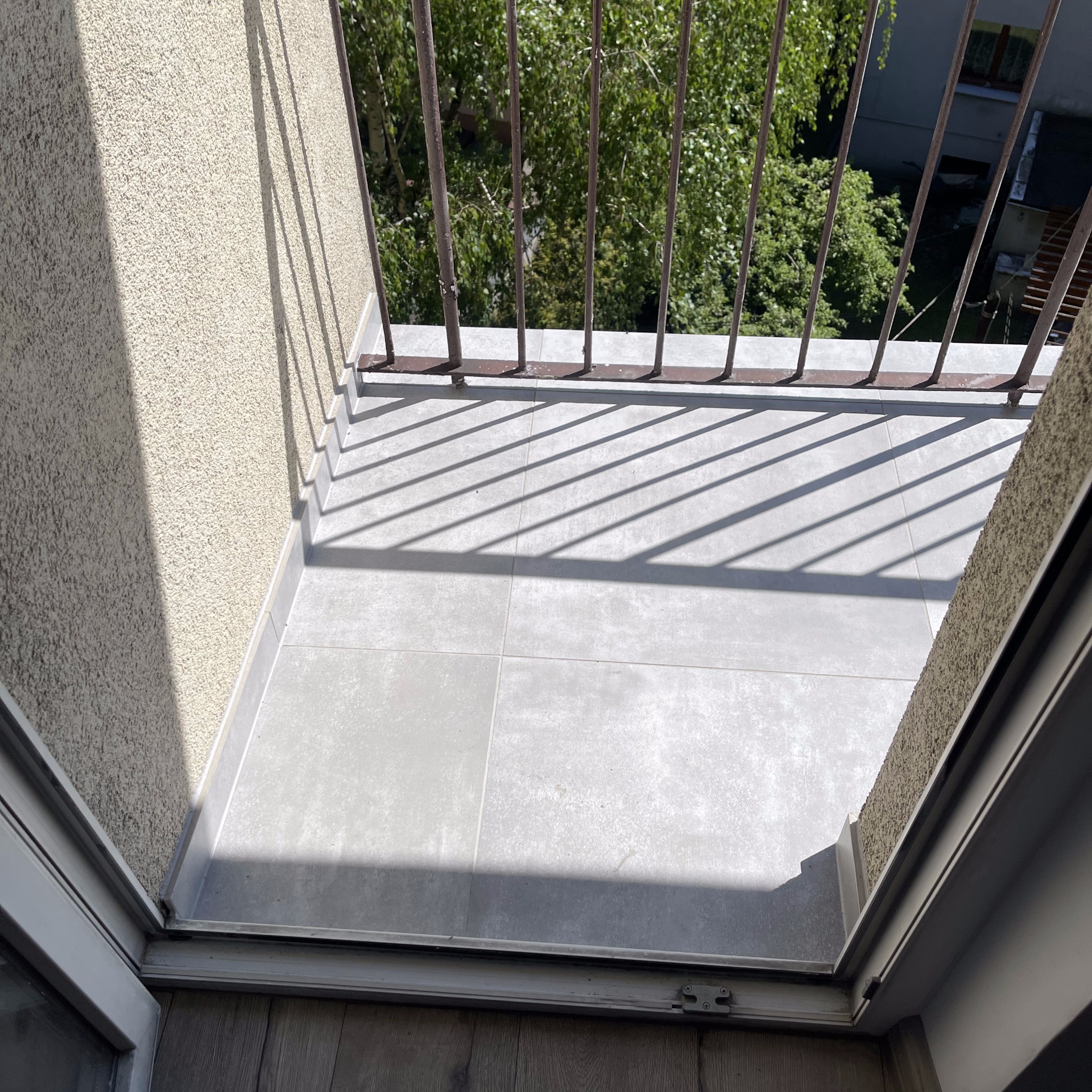 Wykończony balkon w Rybniku z szarymi płytkami podłogowymi i metalową balustradą, widok od strony drzwi balkonowych, z widocznymi cieniami balustrady na podłodze.