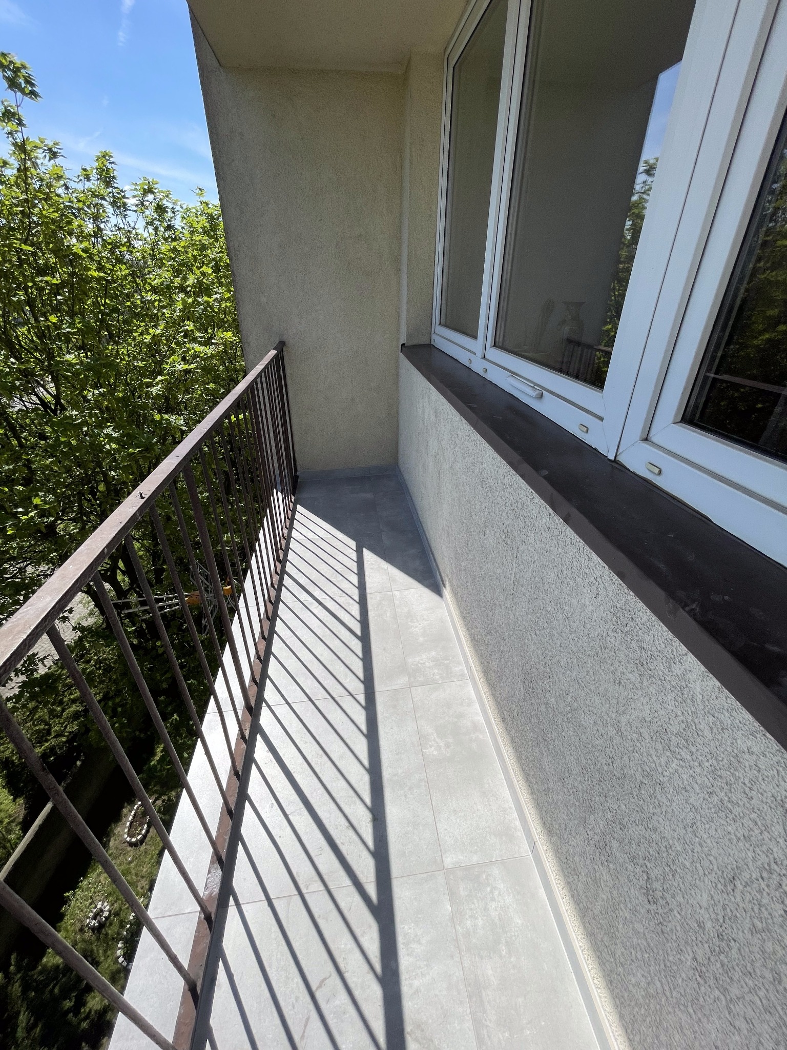 Wykończony balkon z szarymi płytkami podłogowymi, metalową balustradą i oknami PCV, widok z góry na zieleń za balustradą, jasne światło słoneczne tworzy wyraźne cienie.