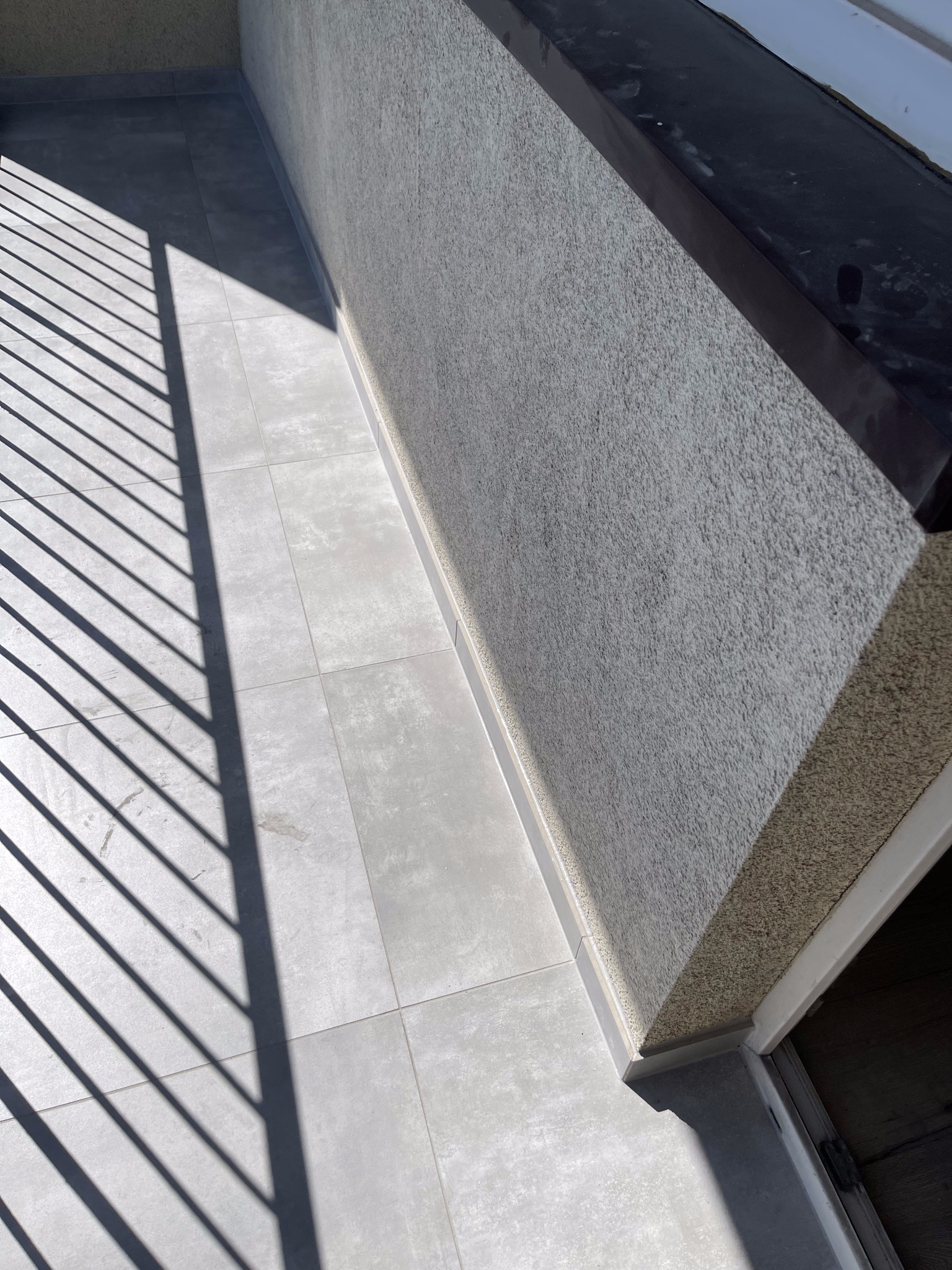 Jasnoszare płytki na balkonie w ostrym świetle słonecznym, cień barierki, ściana z szorstkim tynkiem.