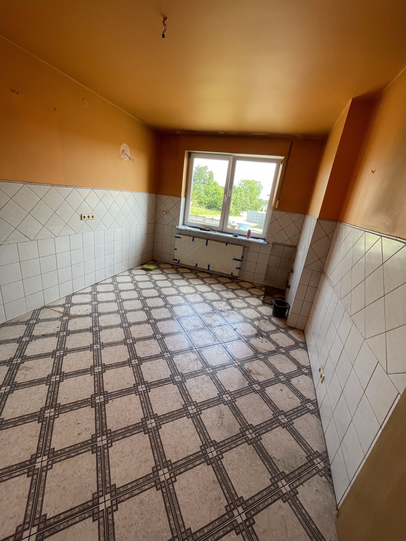 Kuchnia przed remontem: pomarańczowe ściany, białe płytki do połowy wysokości, wzorzysta podłoga, okno z widokiem na zieleń, widoczne ślady użytkowania.