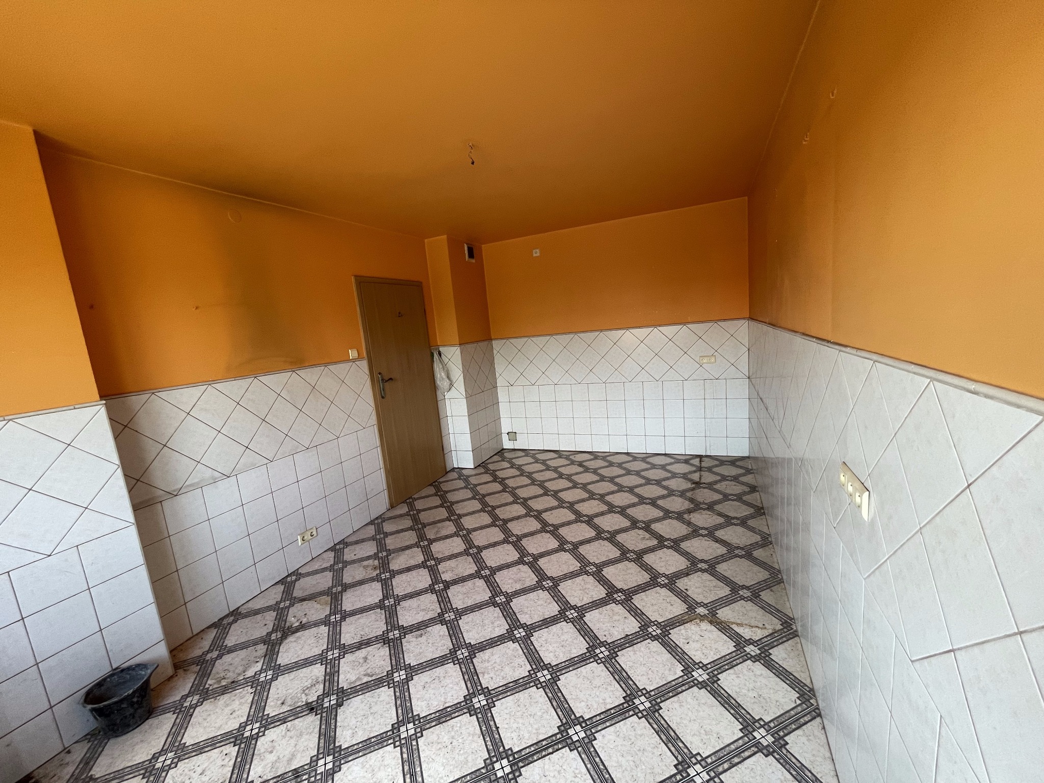 Pomieszczenie w trakcie prac przygotowawczych do remontu: pomarańczowe ściany do połowy wyłożone białymi kaflami, wzorzysta podłoga, widoczny kubeł i drzwi.